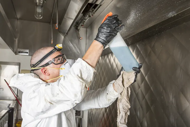 Technician in white coveralls, goggles and a headlamp reaching overhead with a spray bottle and cloth to clean the underside of a stainless steel exhaust canopy, seen from below