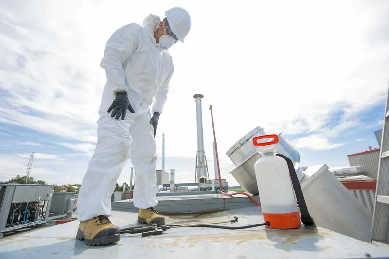 Technician in white coveralls and a hard hat on a flat commercial roof beside a pump sprayer, with an upblast exhaust fan behind him