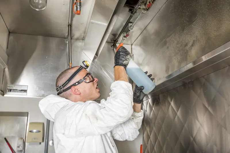 Technician spraying degreaser along a stainless steel kitchen canopy above a commercial cook line