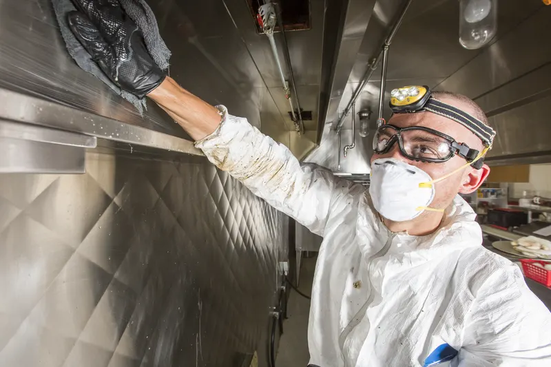 Technician in white coveralls, goggles and a headlamp reaching up with a gloved hand inside a commercial kitchen exhaust hood
