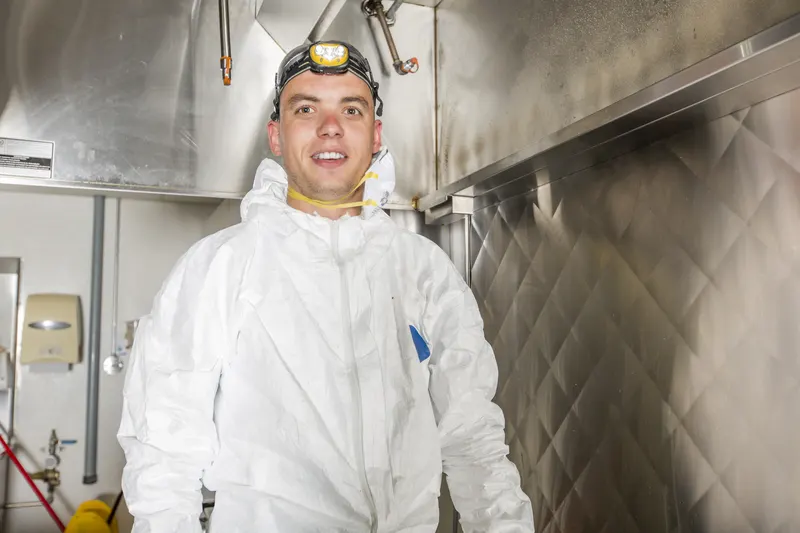 Technician in white coveralls with a headlamp and respirator standing beneath a stainless commercial kitchen hood beside the quilted backsplash