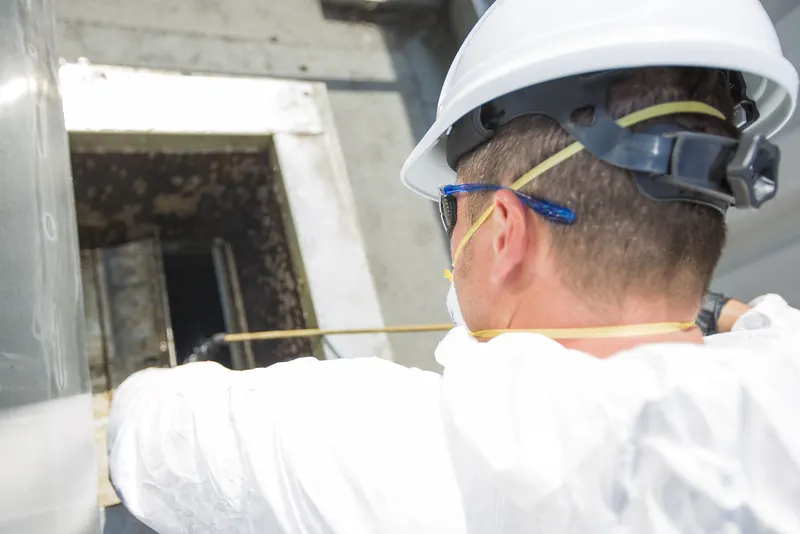 Worker in a hard hat and safety glasses reaching into an open access panel on a grease-darkened commercial exhaust duct