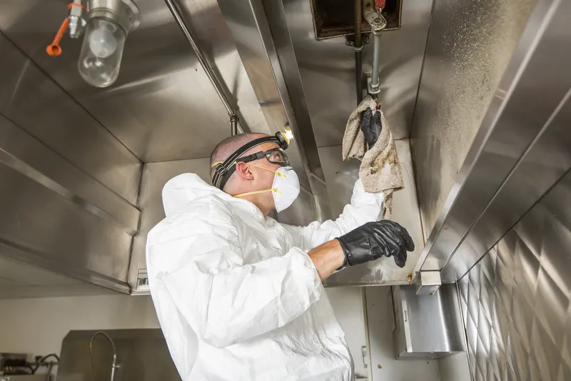 Technician in a protective coverall and respirator wiping the lower edge of a stainless steel exhaust canopy with a cloth