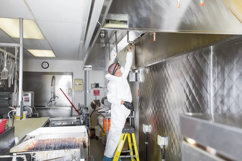 Technician standing on a step ladder wiping the front lip of a stainless steel kitchen exhaust canopy while the cooking equipment below is covered