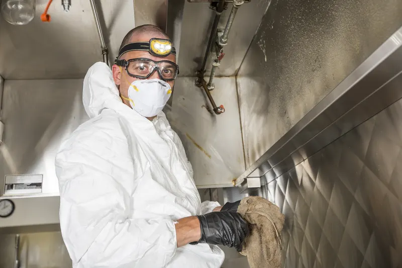 Technician in a white coverall, goggles and headlamp holding a cloth at the filter slot along the front rail of an exhaust hood